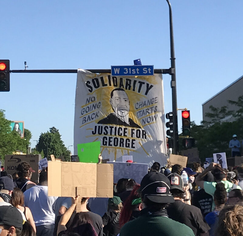 Protestors marching down a street in Minneapolis. A banner hangs off the sign post for W 31st St. The banner shows a drawing of George Floyd and reads "Solidarity," "Justice for George," "No Going Back," and "Change Starts Now."