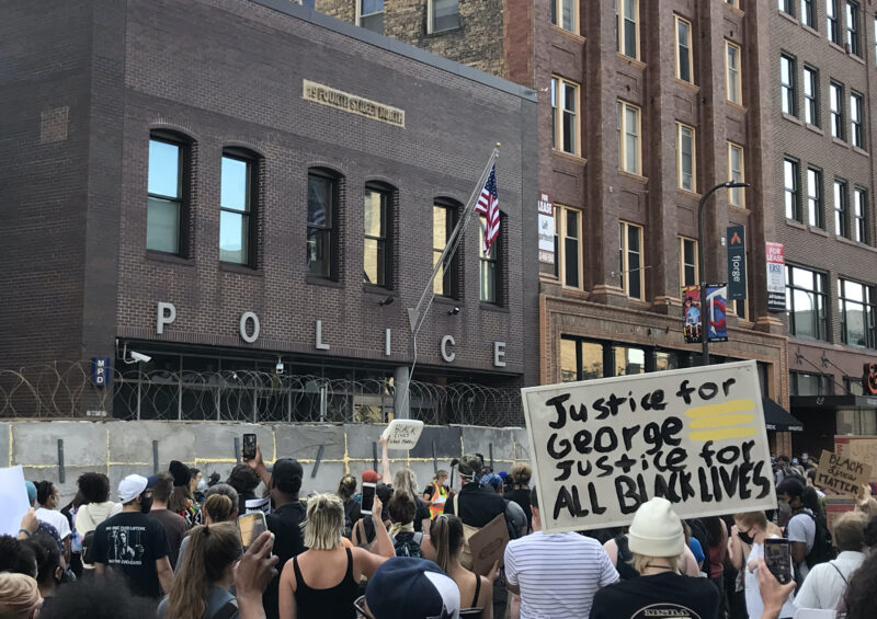 Protestors gather in front of a Minneapolis police precint with signs like one that reads "Justice for George. Justice for All Black Lives." 