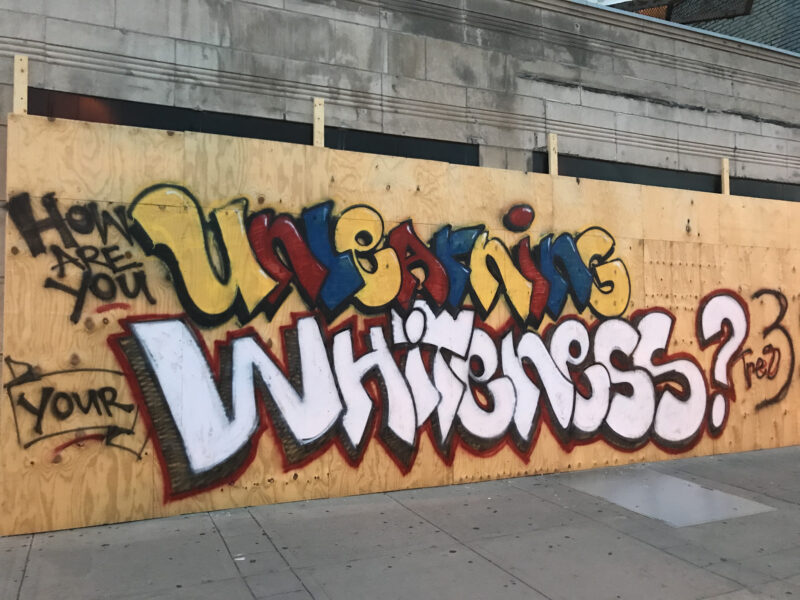 Plywood boarding up a building in Minneapolis. The graffiti on the board reads "How are you unlearning your whiteness?" The "unlearning" is in bold yellow, red, and blue colors. "Whiteness" is in a big, stark white color.