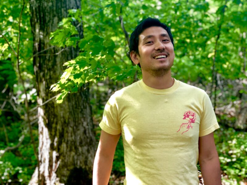 A photo of the author, Khoi Nguyen, wearing a yellow t-shirt while standing in the woods.